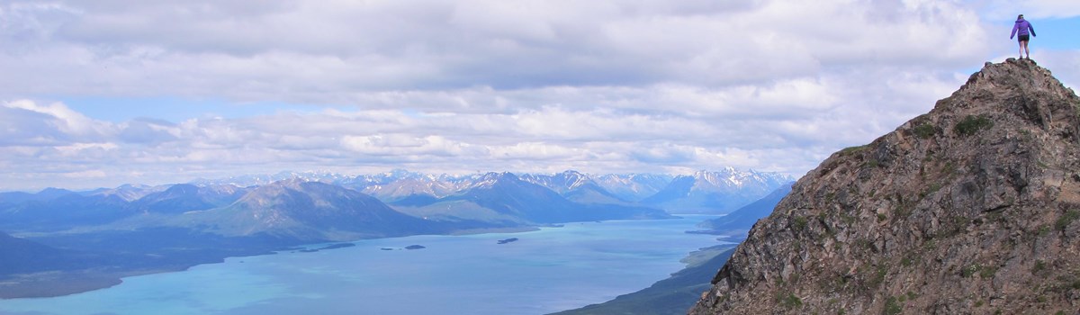 A woman stands on top of a rocky mountain looking out towards the turquoise water of Lake Clark