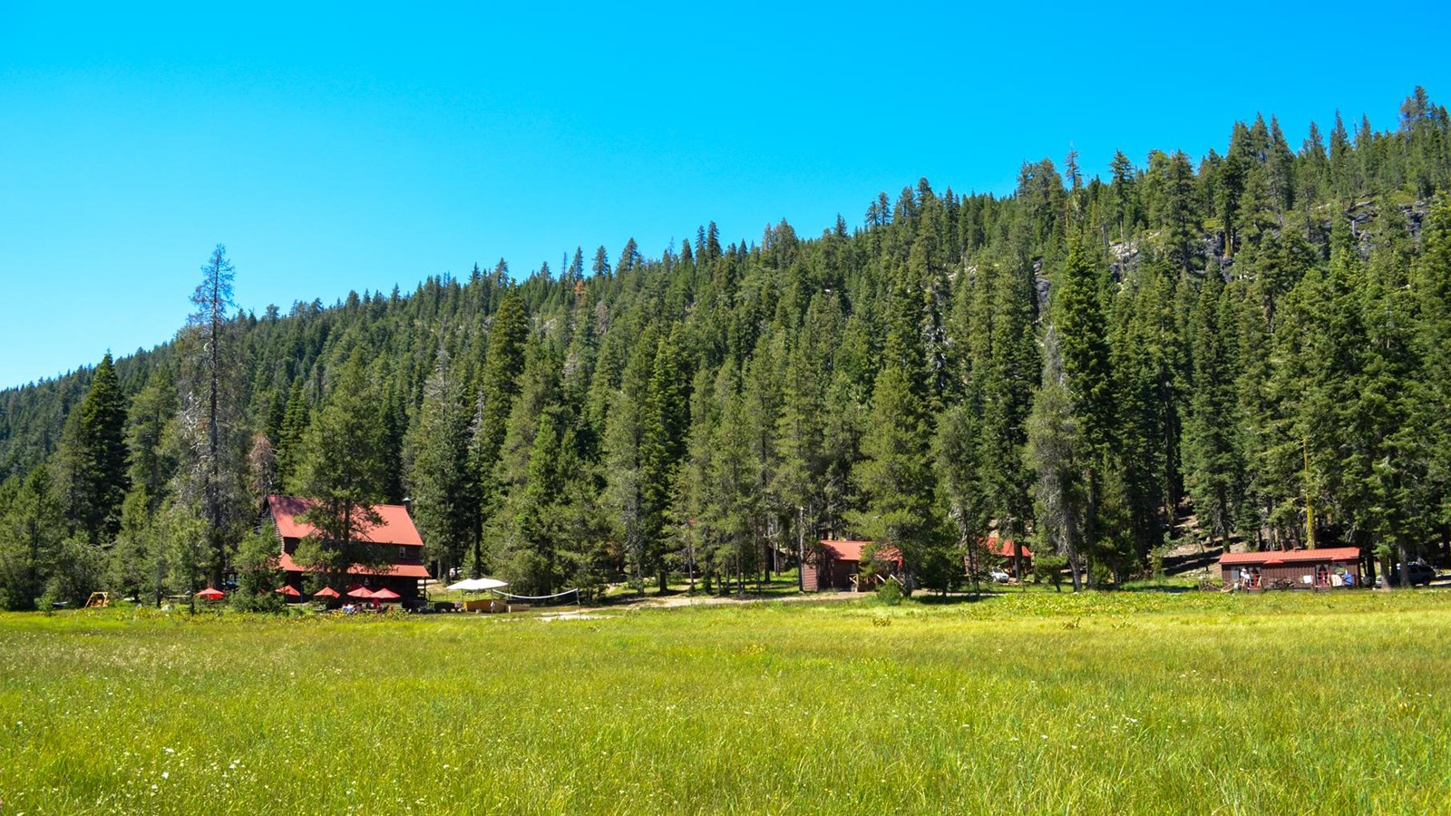 A two-story red and brown lodge and three cabins at the base of a hillside fronted by a meadow. 