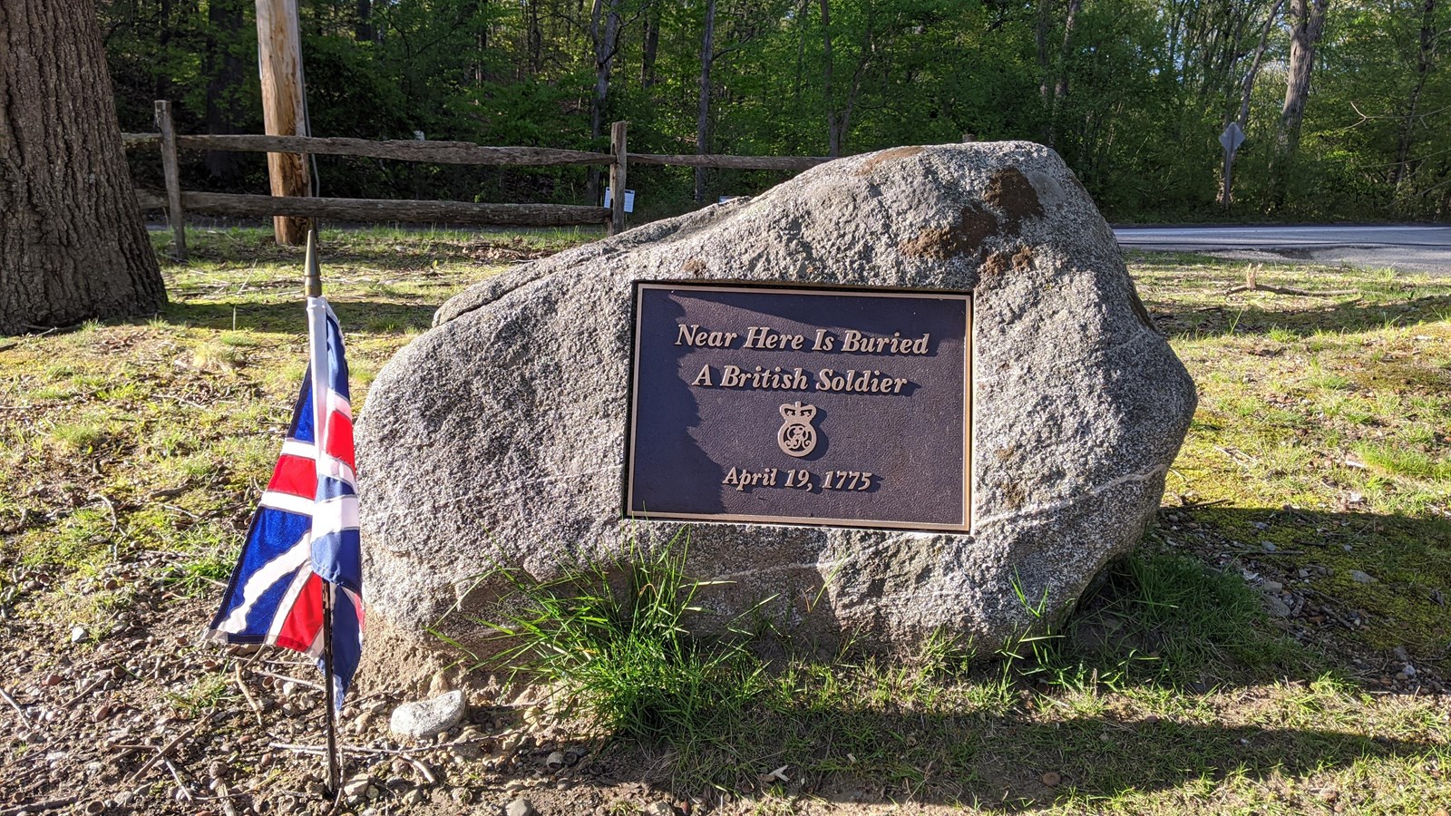 A stone grave marker for a British soldier with metal plaque sits in low grass next to a modern road