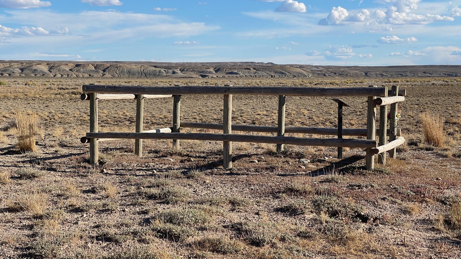 Small fenced grave, sign, stones; barren ground, plateau, blue sky, clouds in sky.