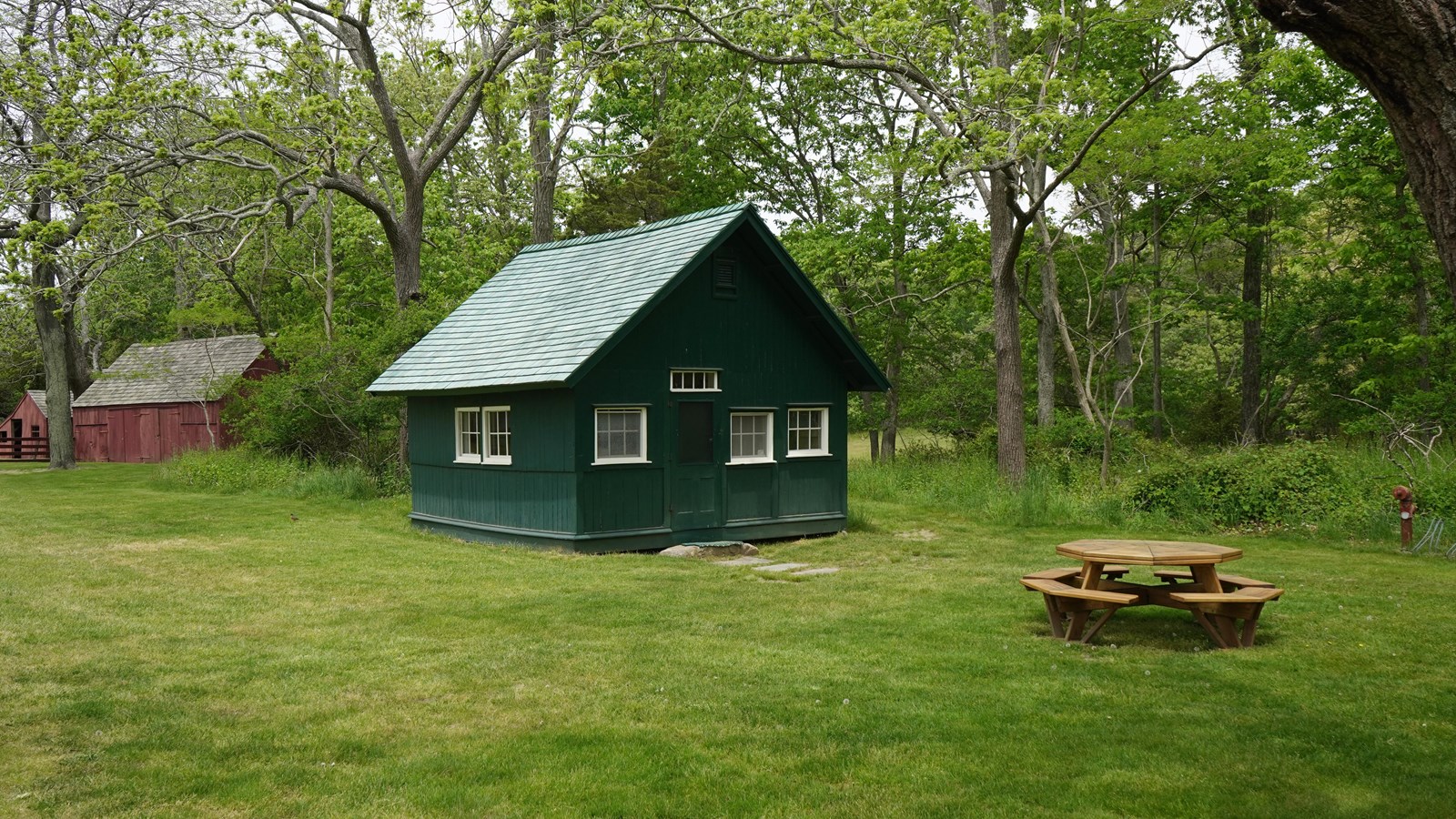 A small green and white building stands next to a picnic table, barn and trees.