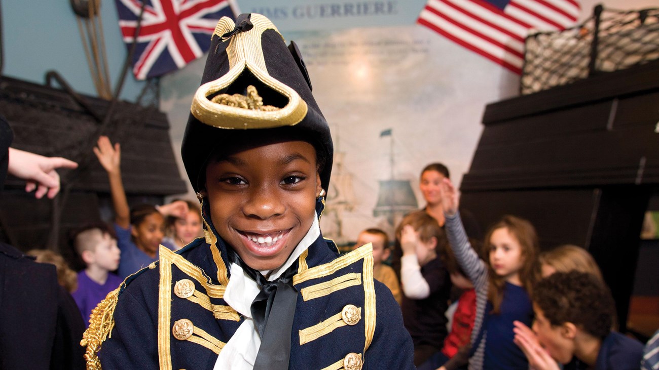 Young girl in a blue 1812 captain\'s uniform while kids learn in background
