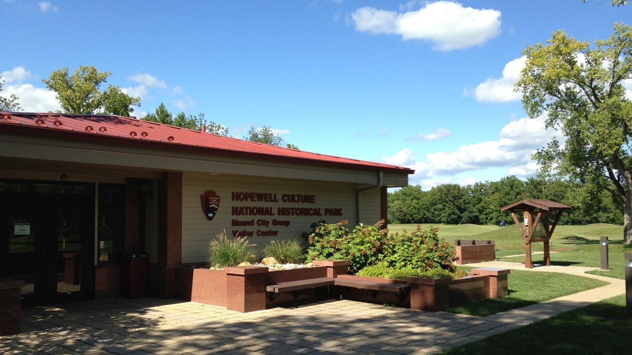 A tan-sided building with red roof in front of green, grass-covered mounds in the background.