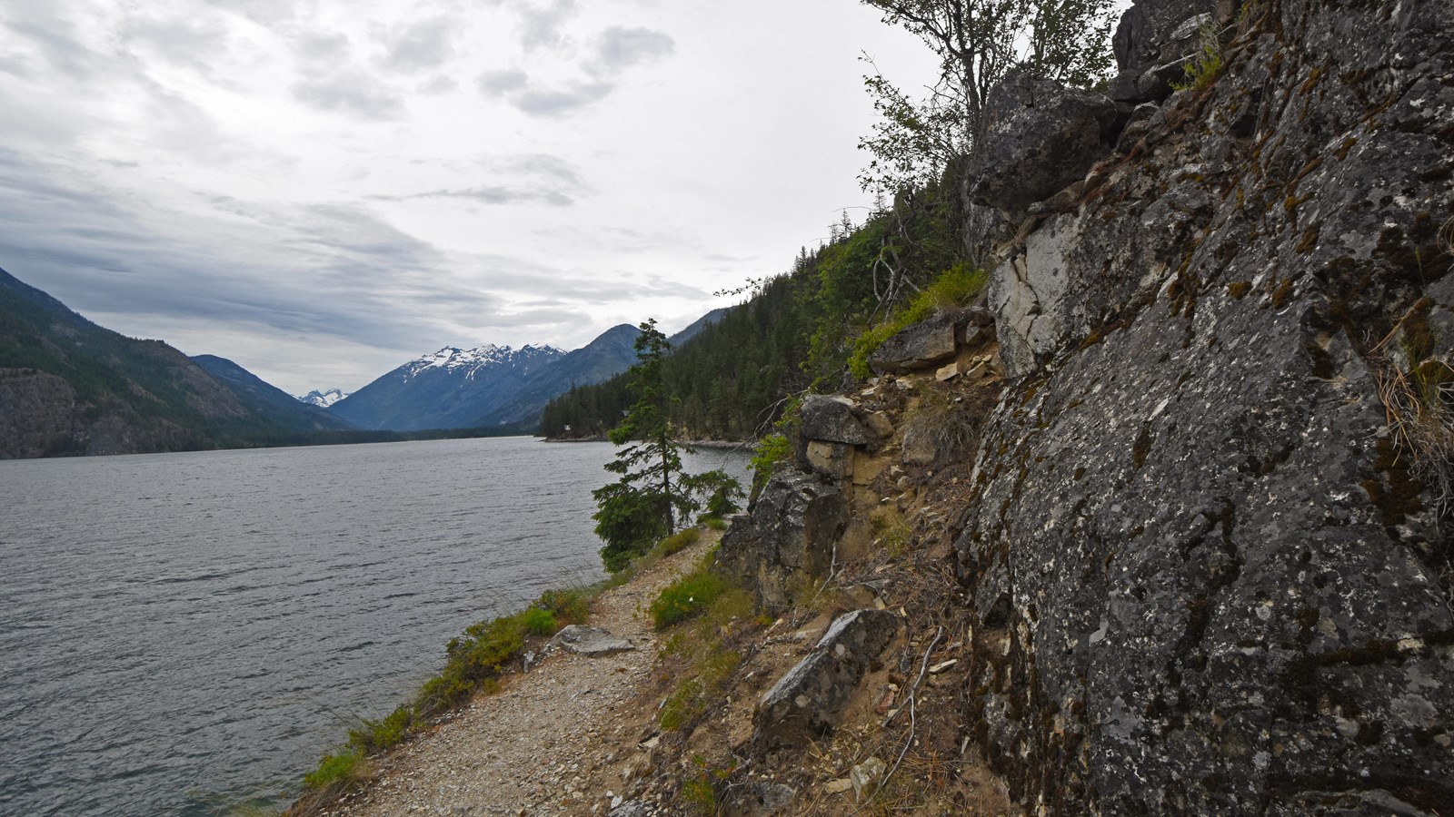 A dirt path follows the shore of a large lake, with snow-capped peaks in the distance.