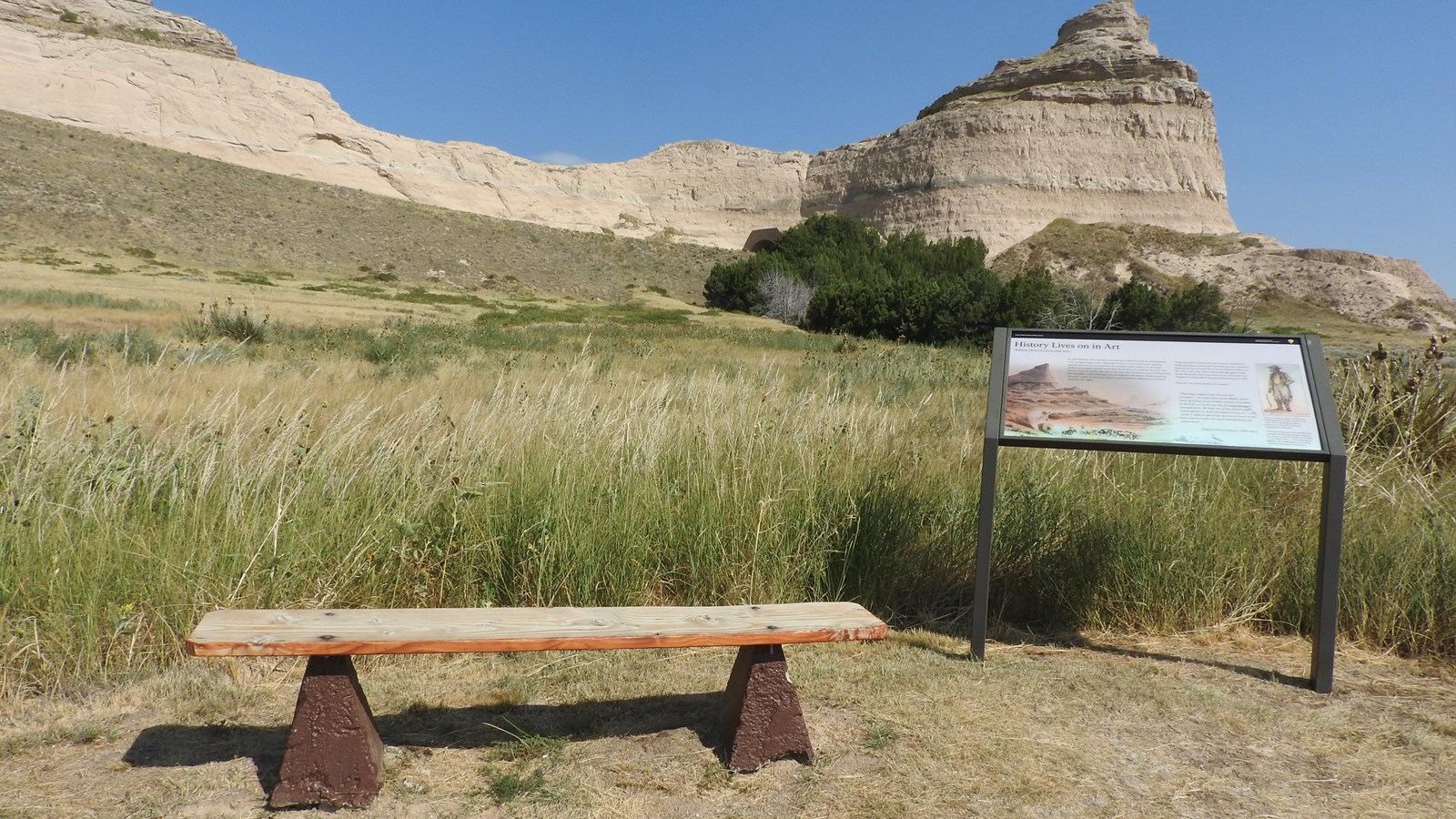 An wayside panel is flanked by a wooden bench to its left. A sandstone bluff is off in the distance.