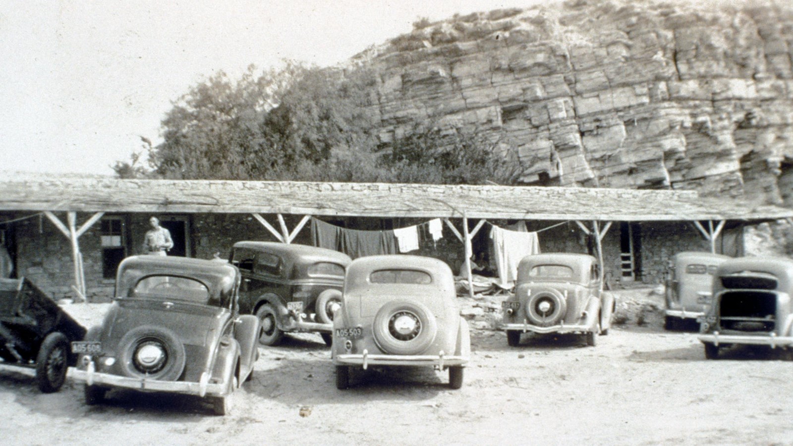 Six cars are parked in front of a long, low stone structure with four visible doorways.