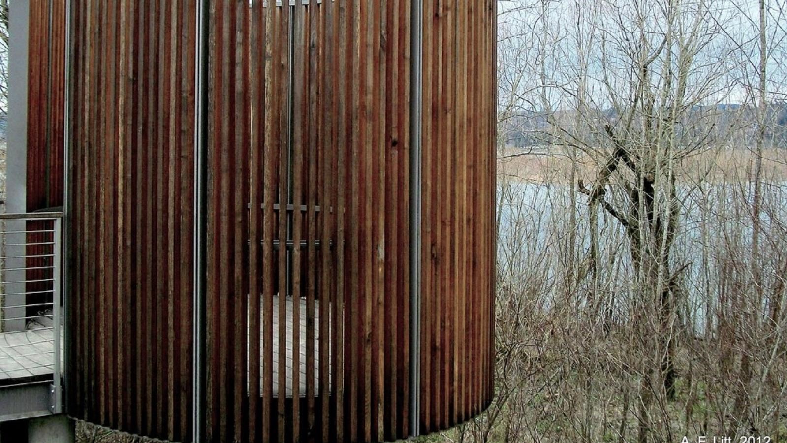 An oval bird blind comprised of vertical wooden slats surrounding an observation platform