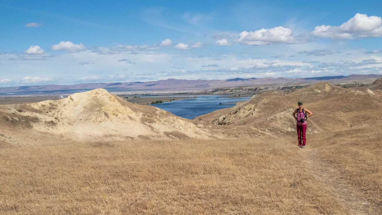 A lone hiker standing on an arid plateau with water and mountains in the distance.