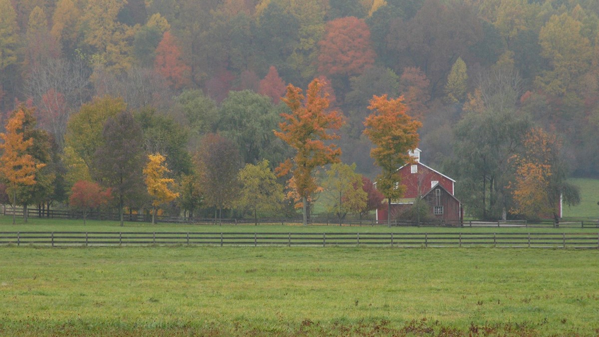 Scenic view of red barn with grassy fields in foreground and hillside of colorful leaves behind. 