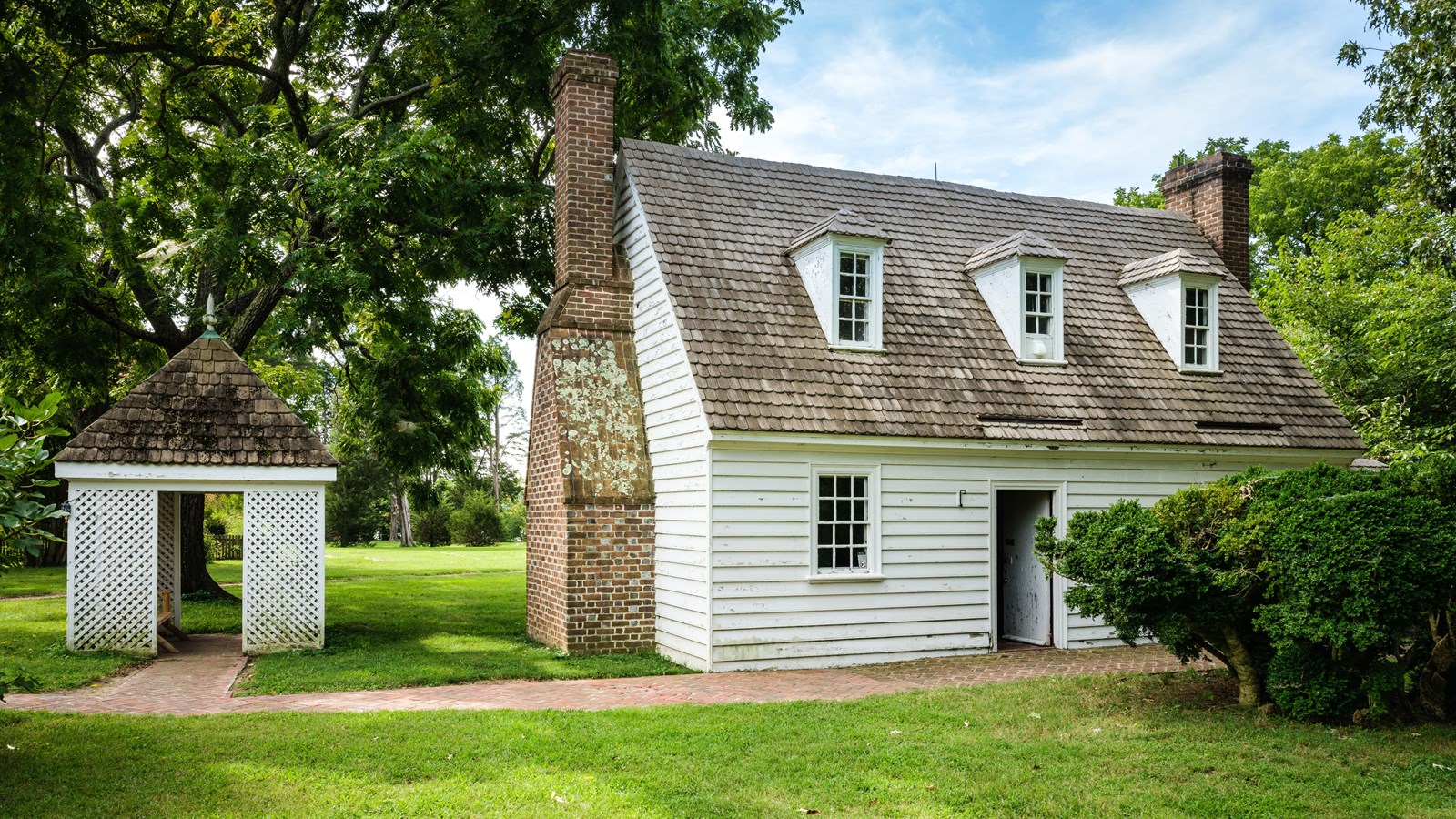 A white building with a door open, a green bush in front, and a brick path.