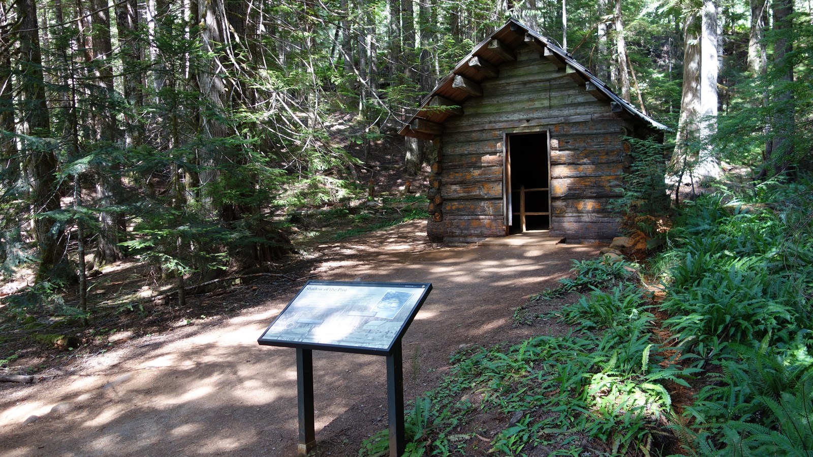 A free-standing metal exhibit panel stands in front of a wood cabin surrounded by forest.