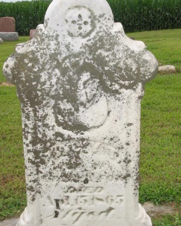 Large headstone with grey spots and faint lettering on it. The top is ornately shaped. 