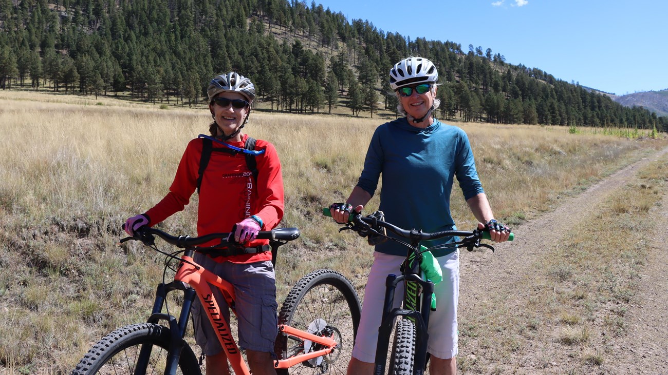 Two people sitting on their bicycles along a rough dirt trail with mountains in the background.
