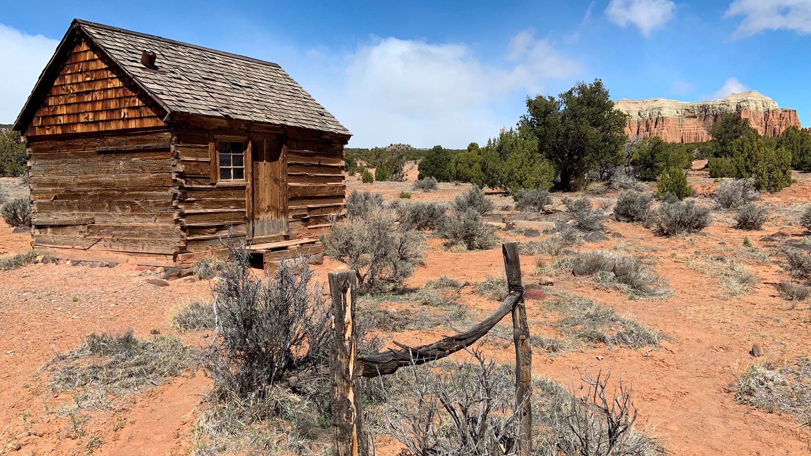 Small, one-room log cabin with wood shingle roof, hitching rack, and colorful rock formations.