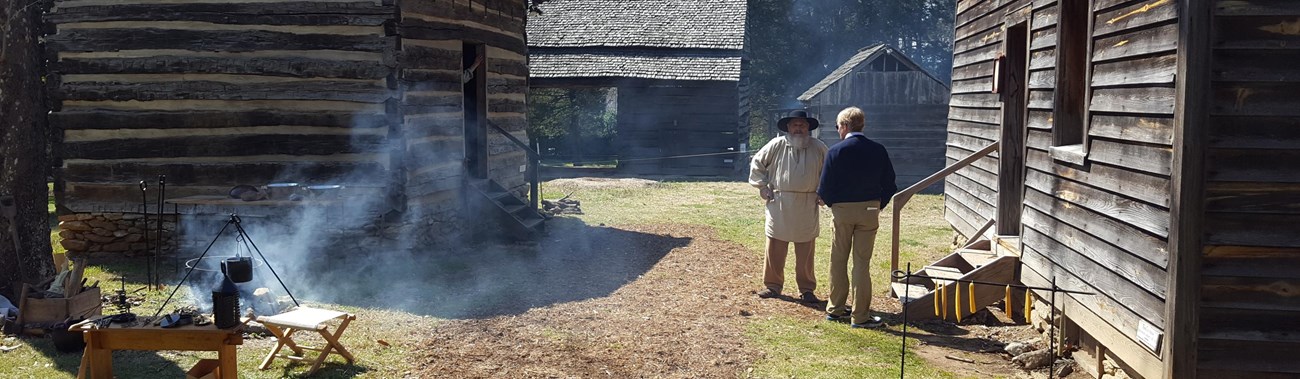 Living history interpreter stands at historic farm with an outdoor fire. 