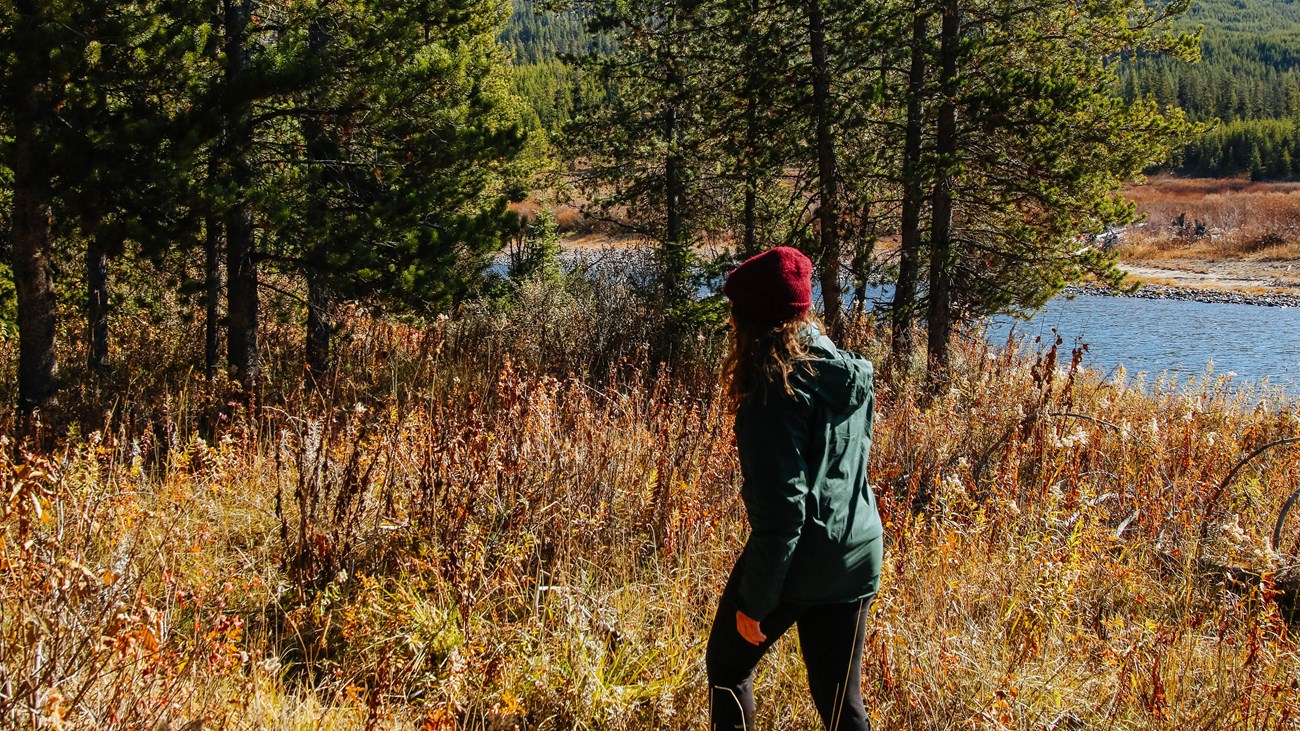 A female hiker walks along a wooded trail by a river.