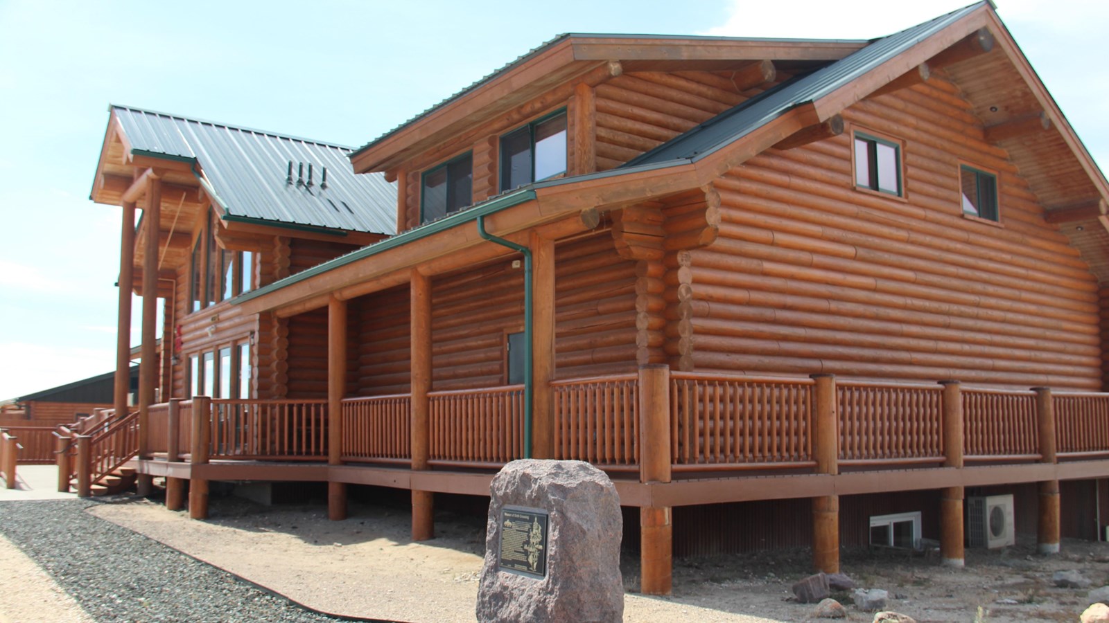 A log cabin visitor center sits with a green roof, wrap around porch, and a monument next to it.