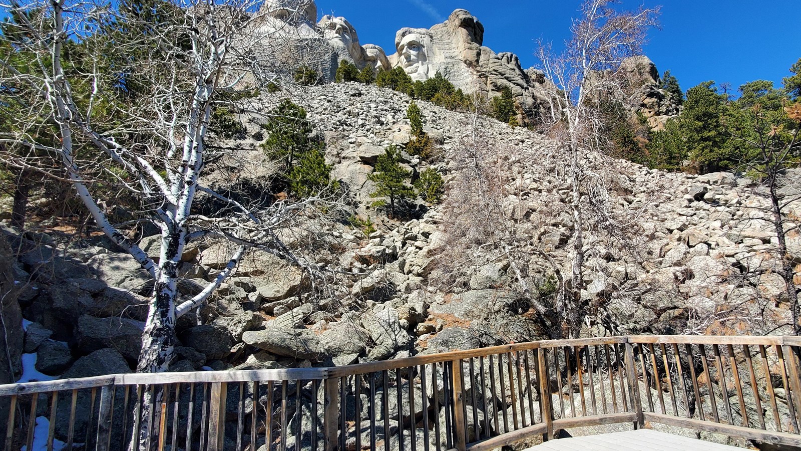 Photo of a wooden terrace with a slope covered in granite rubble and Mount Rushmore near the top.