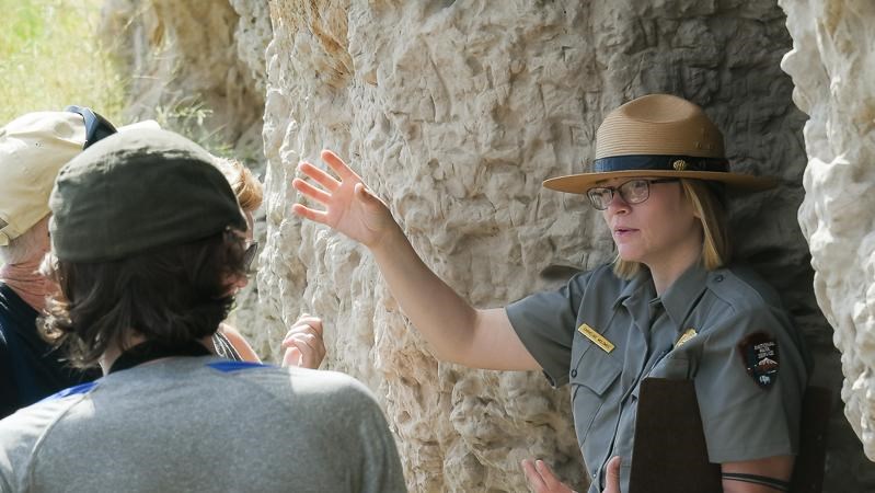 A park ranger talks near a rock outcropping while a person intently watches her.