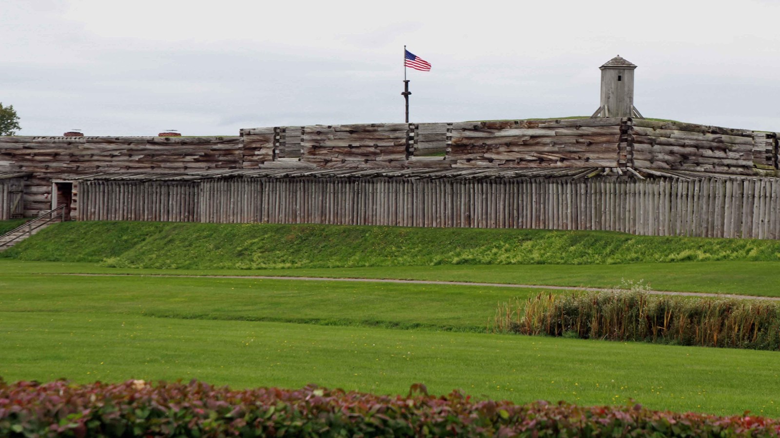 A wide view of the exterior wall of the fort. Big wooden logs with a door and staircase out.