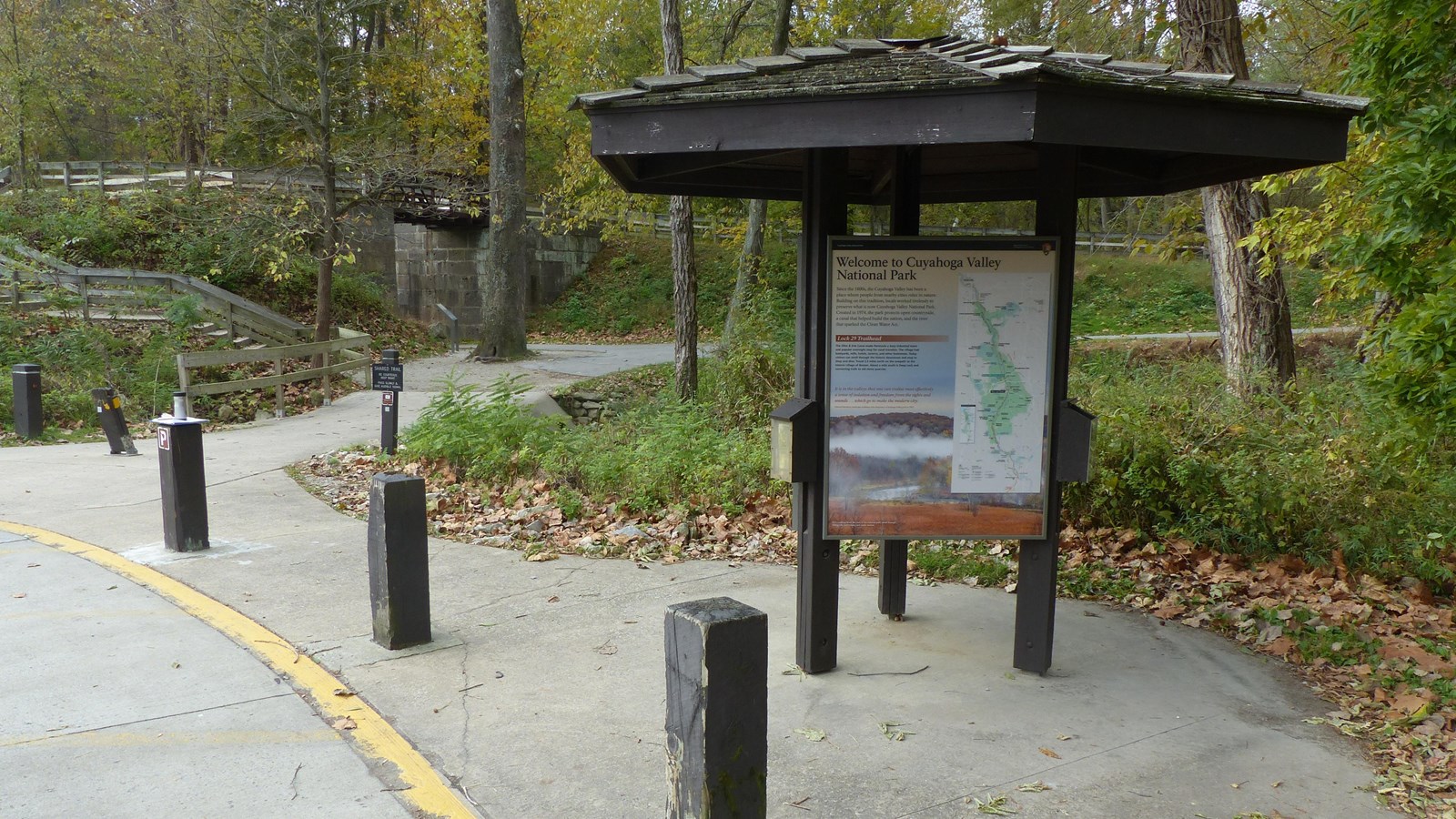 Three sided kiosk beside parking lot posts. A paved path leads to a stone lock and wooded trail.