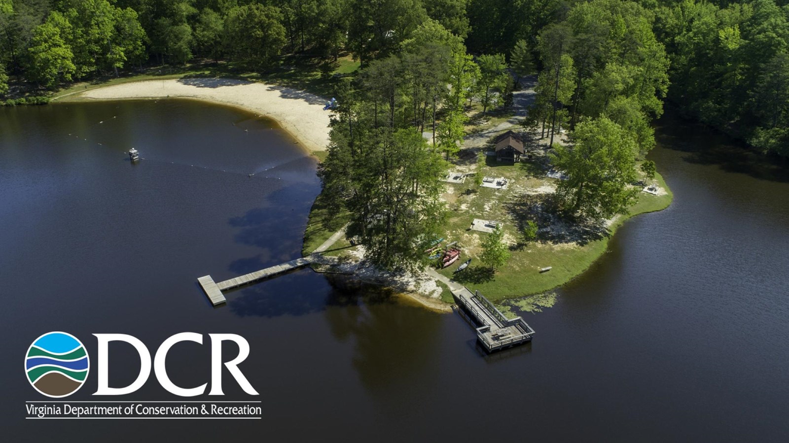 Aerial view of a lakeside area featuring a sandy beach, wooden dock surrounded by green trees.