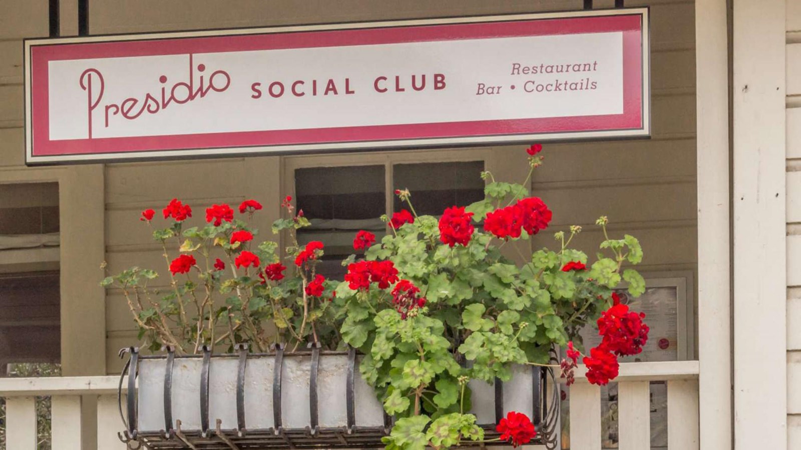 Presidio Social Club sign and bright red geraniums on front porch.