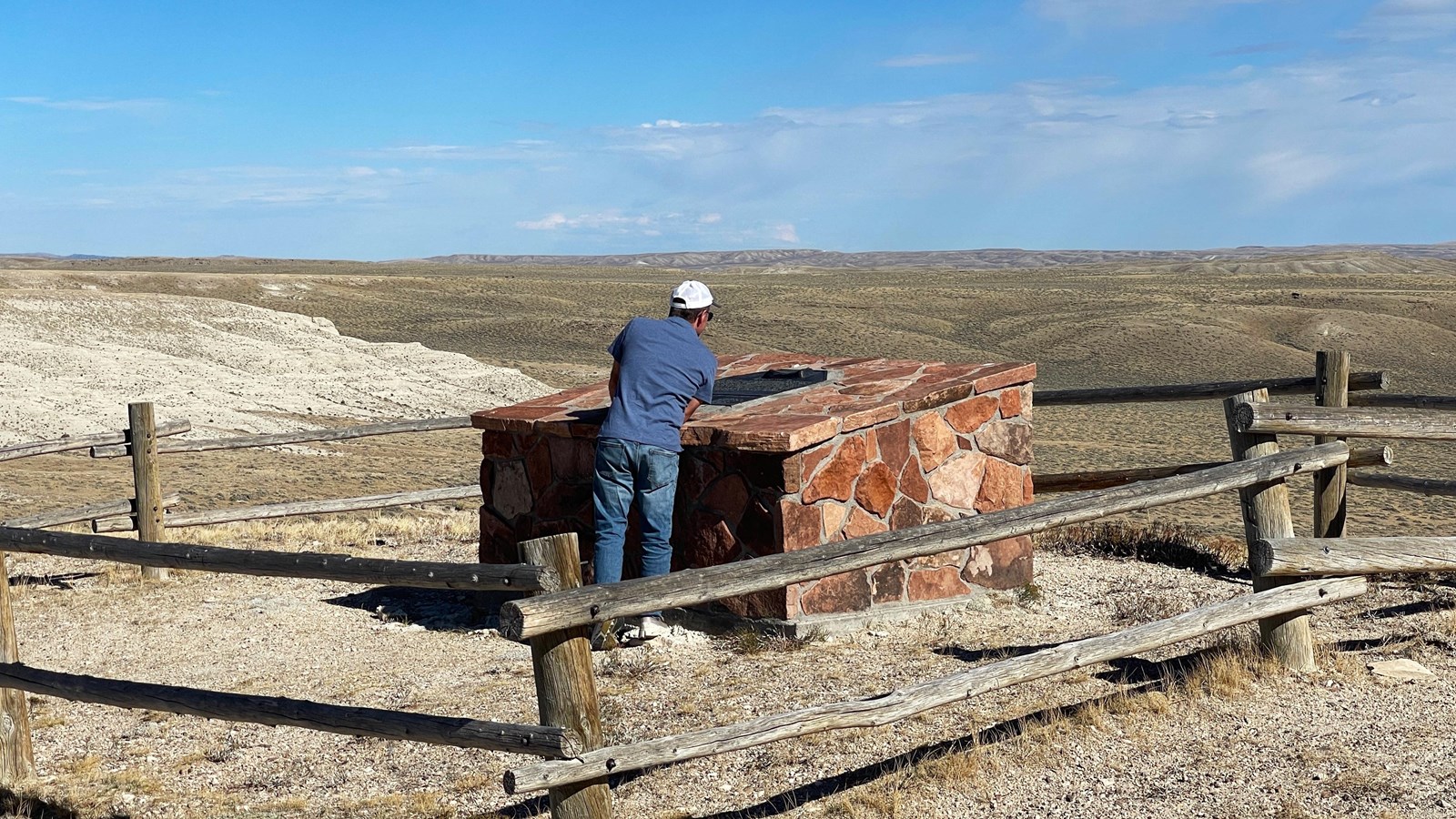 A man reads plaque on red stone monument; arid landscape, blue sky, gray clouds above.