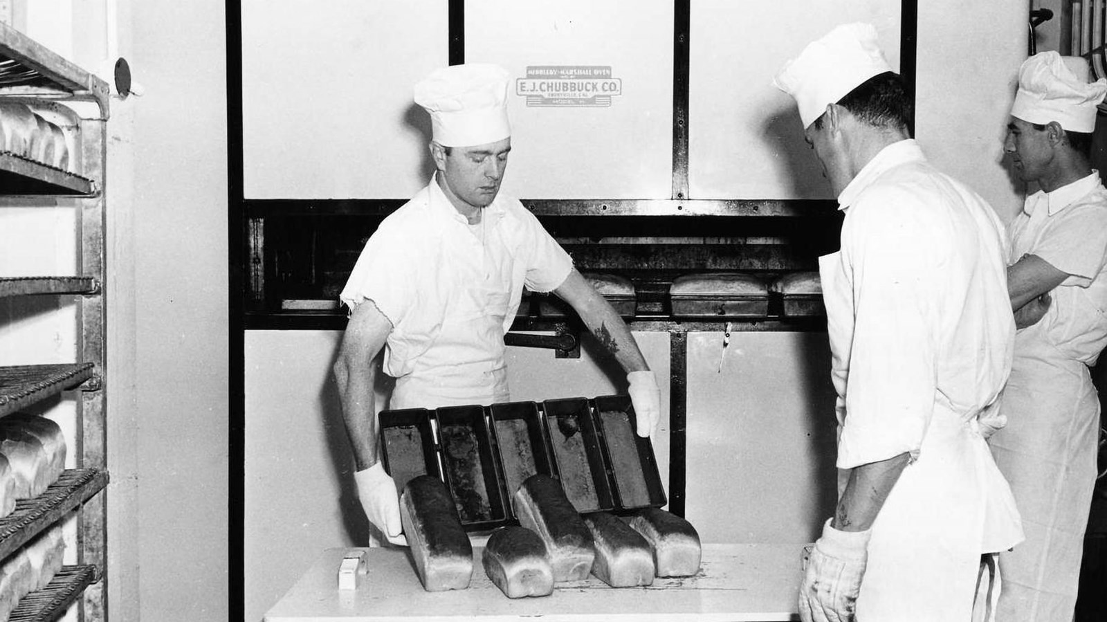Three men stand in front of a commercial oven. One man dumps five loafs of bread onto a table.