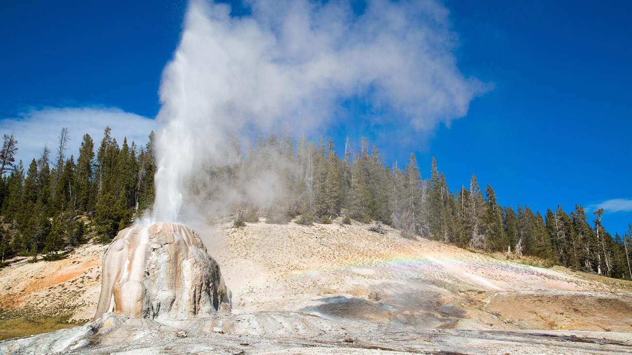 Steam and water erupt from the tan cone of Lone Star Geyser. A rainbow appears in the mist.