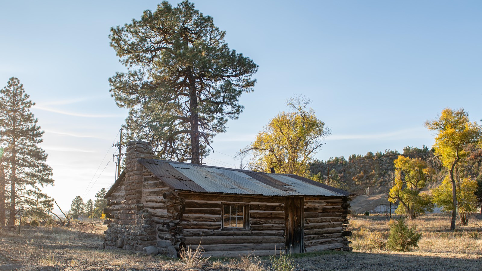 A log cabin sits in a field with trees and hills in the background