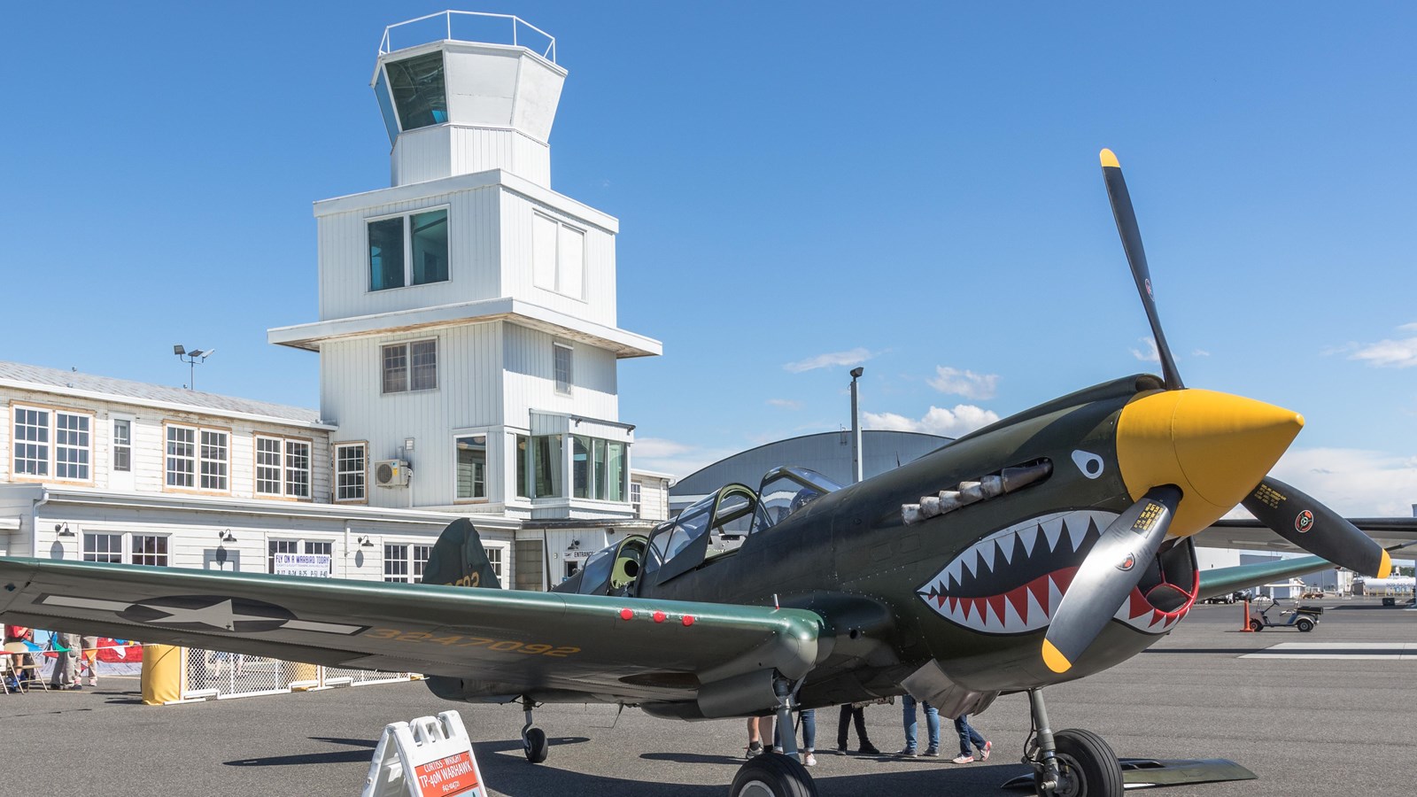 A green military plane is parked in front of a building with a 4 story square observation tower. 