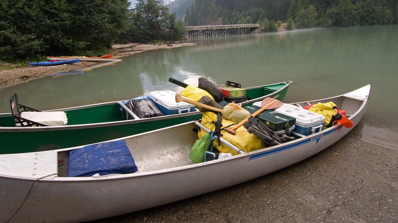 Two silver canoes loaded with camping gear sit on the shore of a lake.
