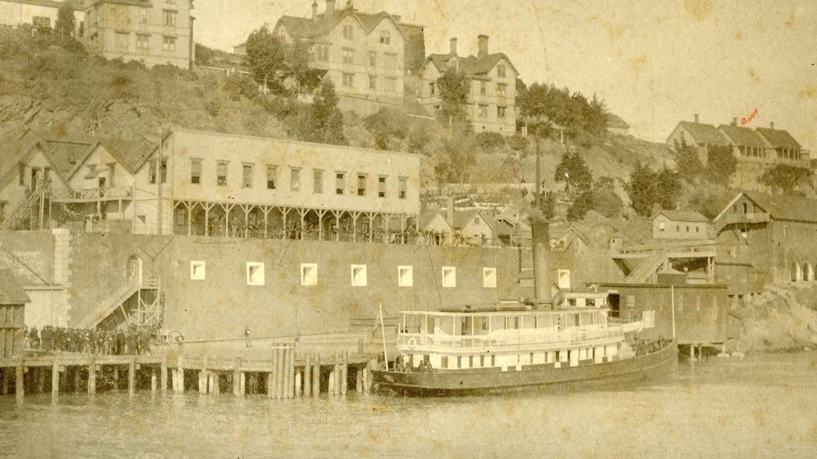 Steamship sits at dock. Large group of men gather to the left of it on pier.