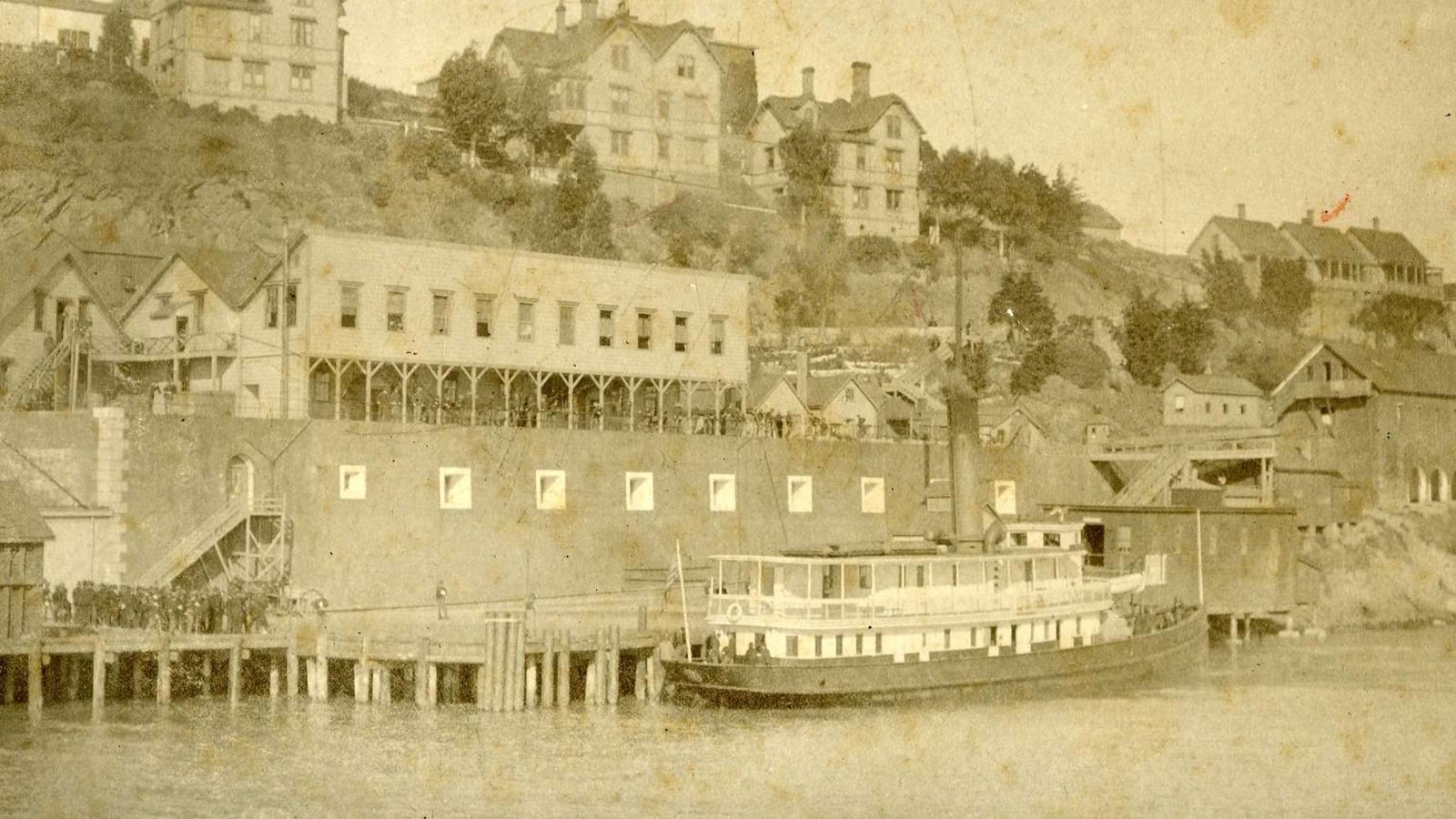 Steamship sits at dock. Large group of men gather to the left of it on pier. 