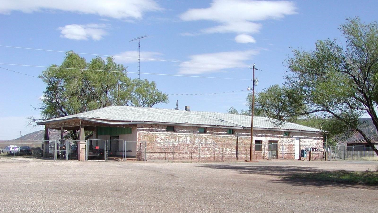 A red brick building with a white roof surrounded by green trees and a gravel parking area.