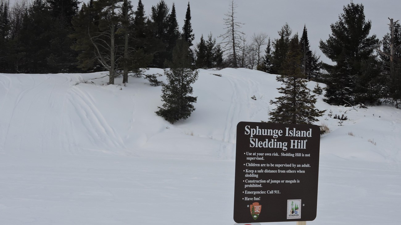 Brown sign titled \'Sphunge Island Sledding Hill\' in front of a hill covered with snow.