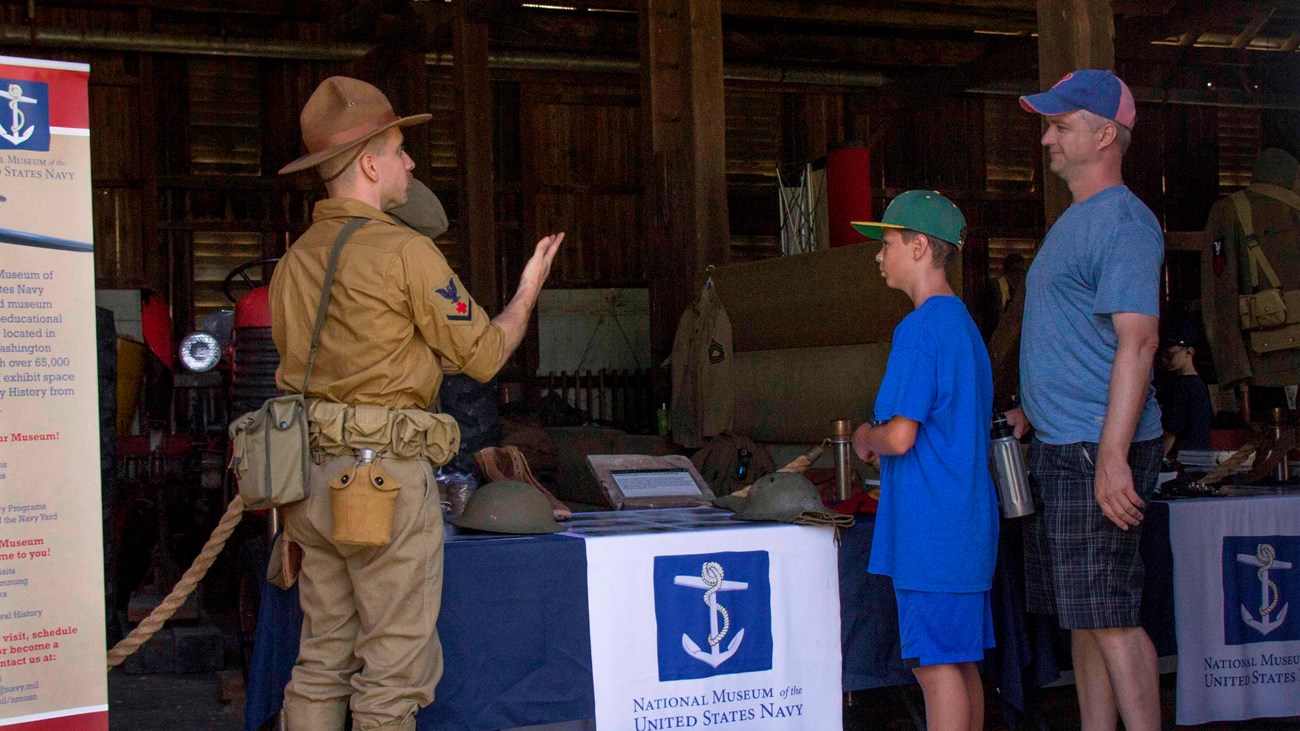 A man in a brown military uniform speaks to two visitors at an interactive table in a barn