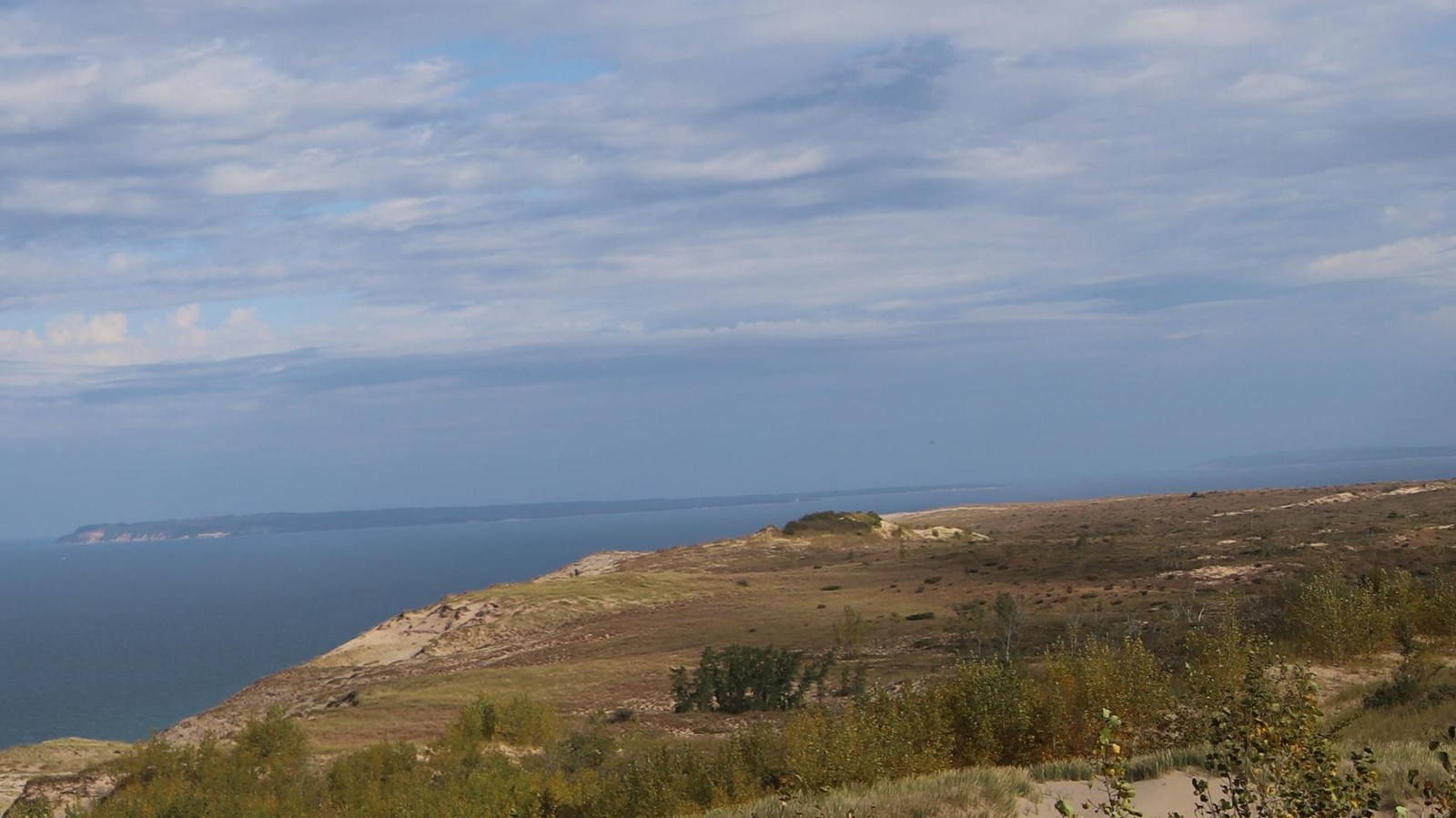 Dunes covered in brown brush greet you. In the distance, a taller eroding dune along the lakeshore. 