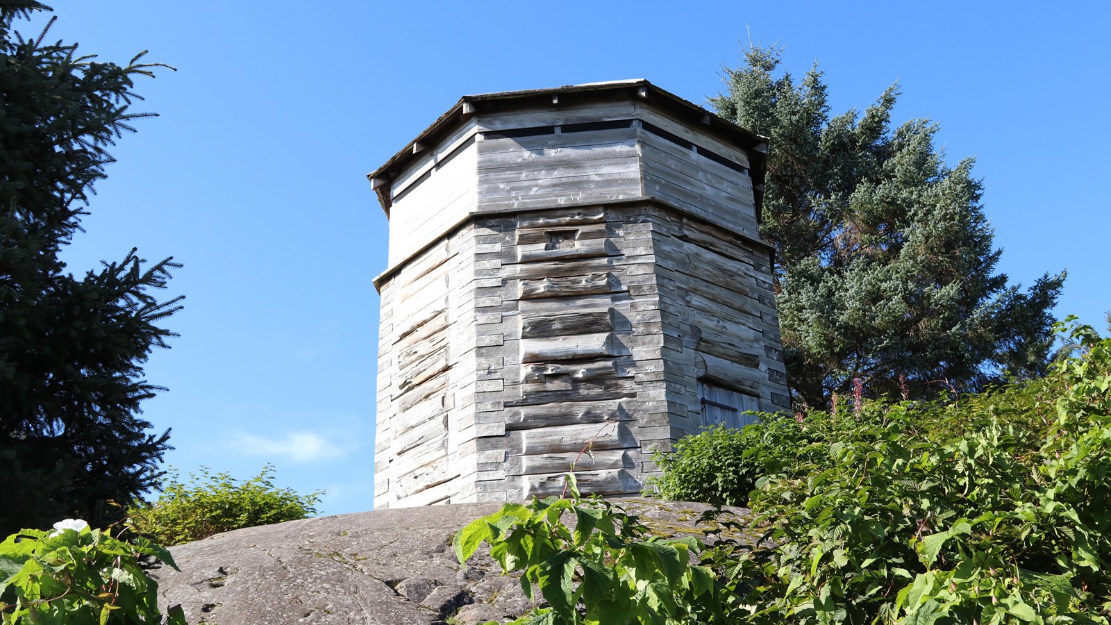 A historic octagonal weathered wooden structure an a rock foundation surrounded by trees.