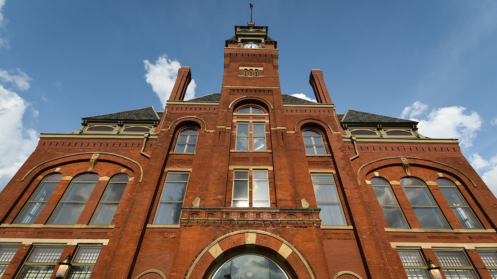 A red brick four story building with a clock tower. Blue clouds behind and reflected in windows.