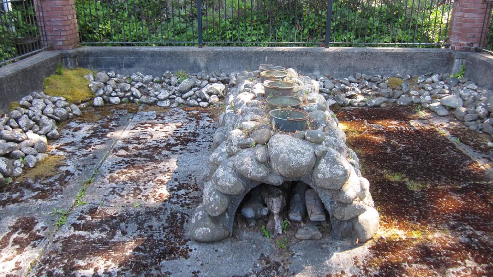 A stone cairn with 5 metal buckets on top sits in the center of small courtyard surrounded by fence