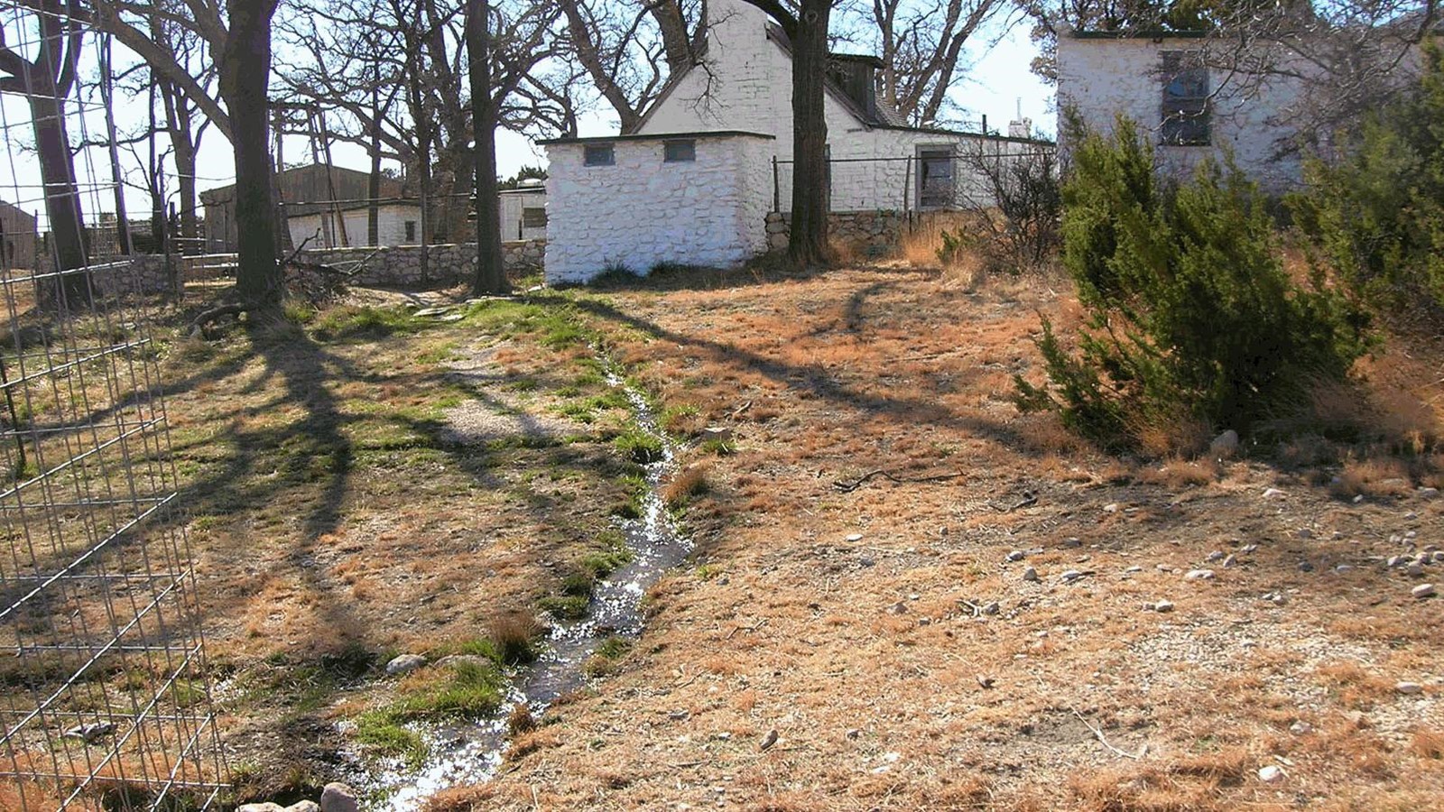 Water flows downslope in a ditch toward a fenced area, left. Tall leafless trees grow around a house