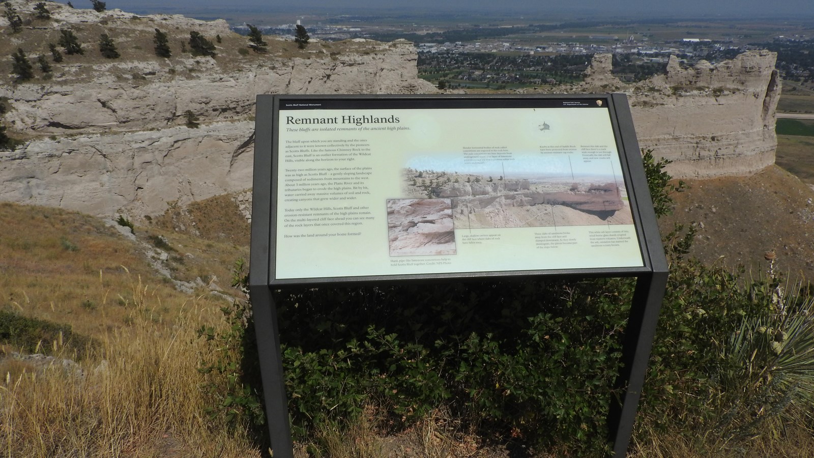 A wayside, interpretive panel sits east of an asphalt-paved trail overlooking a sandstone cliff.