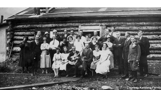 Around two dozen men and women standing in front of a log cabin, posing for a photo