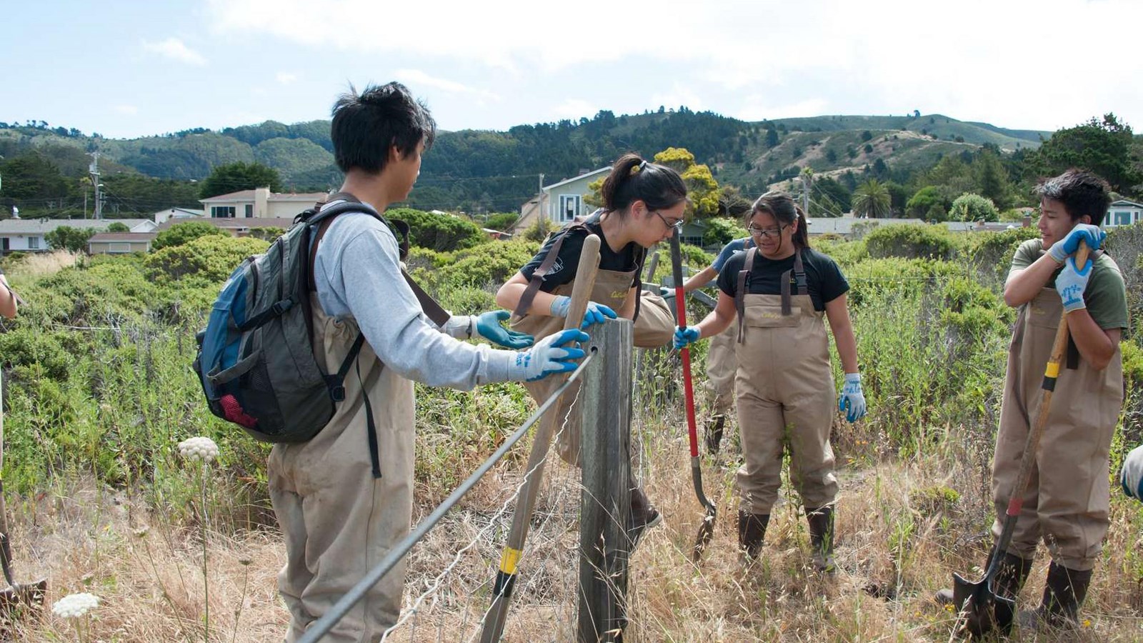 Youth volunteers dressed in waders stand against a fence with shovels.