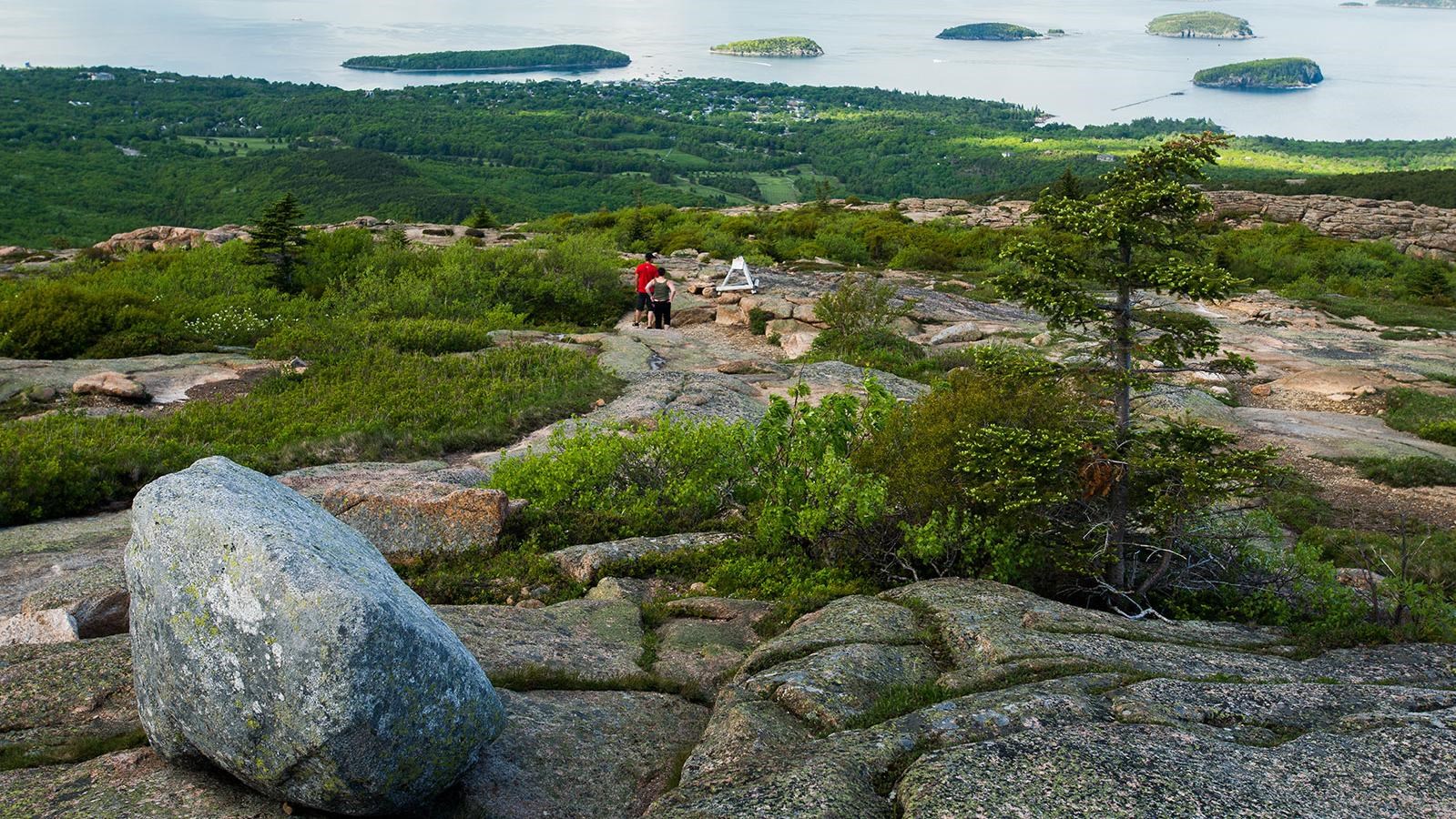 a round glacial erratic sits on a rocky summit with people hiking down the mountain beyond