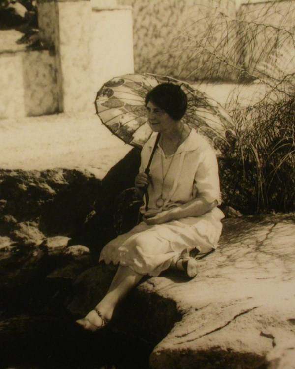 Woman sits beside pool of water with umbrella over her shoulder. 