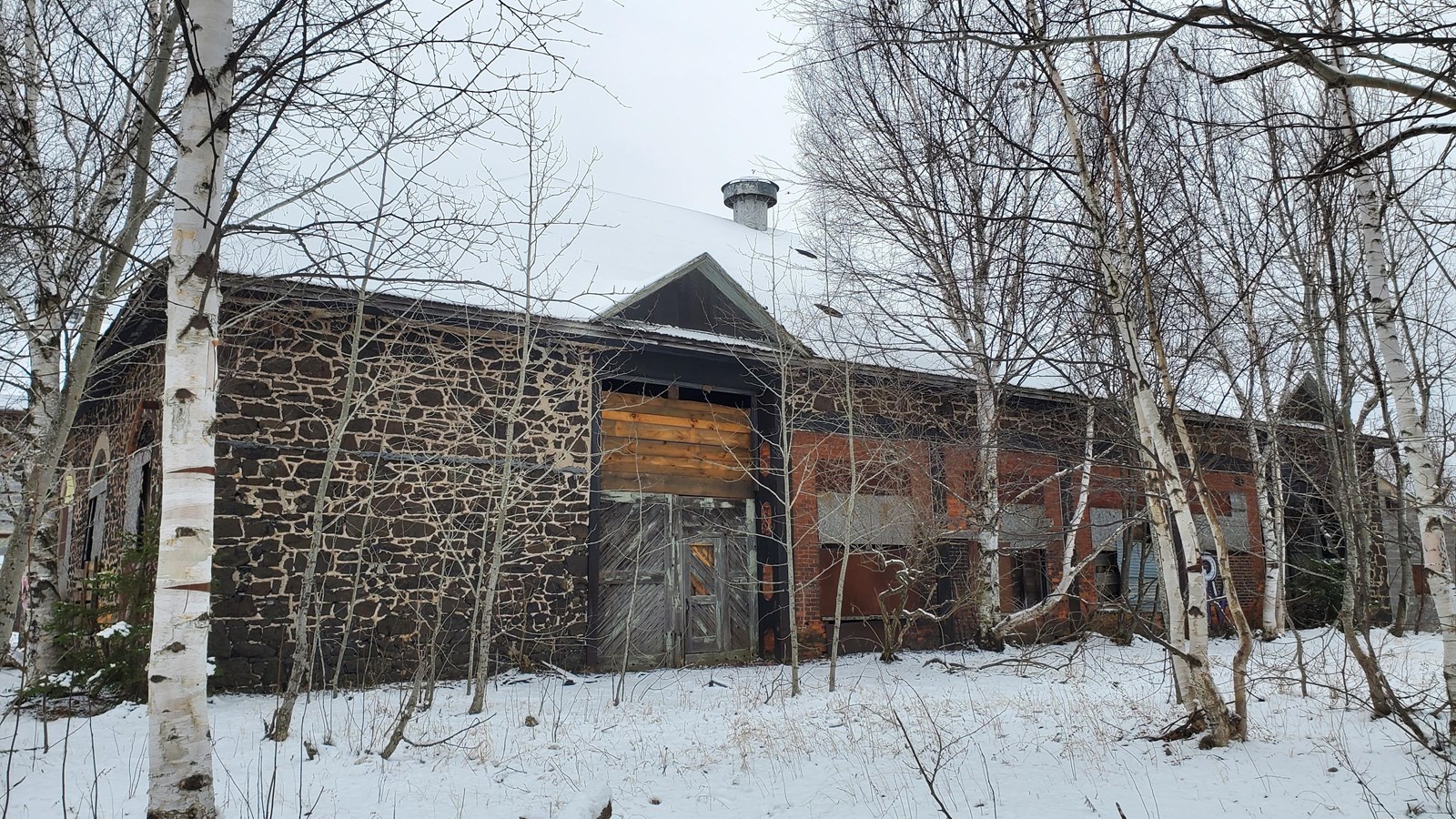 Winter scene of wood and poor-rock building with a large entrance, now boarded and sealed.