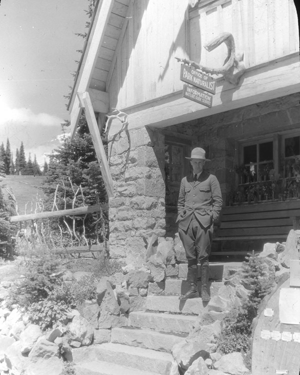 A black and white photo of a ranger wearing a uniform standing on the steps of a historic building.